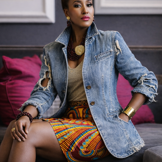 A woman sits on a dark sofa wearing a light-wash distressed denim jacket over a beige top and a colorful orange, yellow, and blue patterned skirt. The background features a dark gray wall with framed art and magenta pillows.